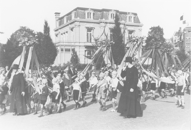 Schwarzes und weißes Foto einer Gruppe von Menschen, die eine Straße entlanggehen und Fahnen halten, mit Bäumen, Gebäuden und einem Maibaum im Hintergrund.