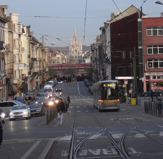 Eine belebte Stadtstraße mit mehreren Fahrzeugen, Fußgängern auf dem Gehweg und Gebäuden auf beiden Seiten, mit einer Tram, einer fernen Brücke, Bäumen und einem klaren blauen Himmel.