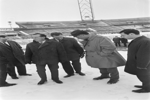 Eine Gruppe von Männern in Mänteln und Schuhen steht zusammen im Schnee, mit einem Stadion, Turm und klarem Himmel im Hintergrund.