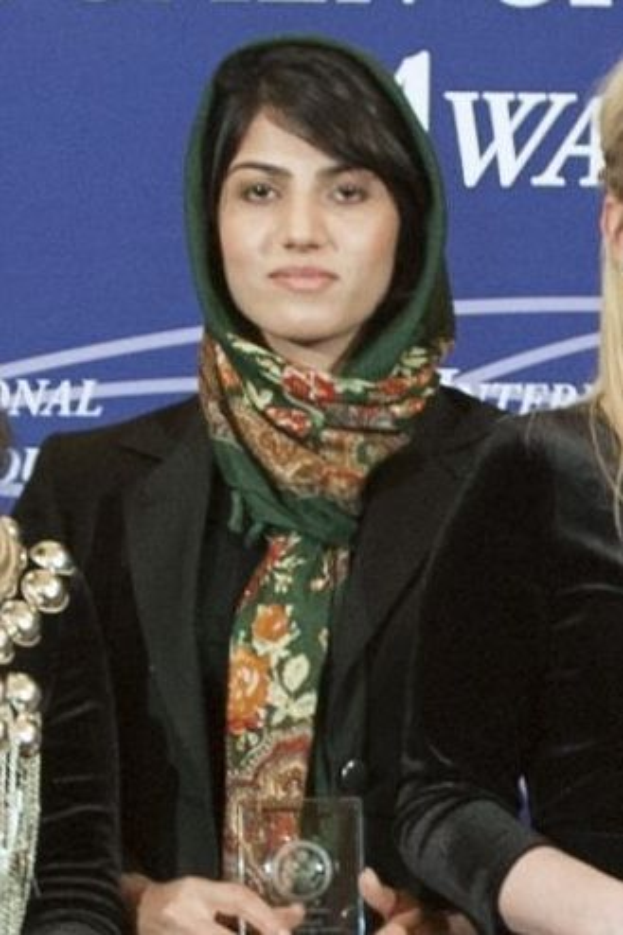 Malala Yousafzai, eine Nobelpreisträgerin für den Frieden, steht in der Mitte in einem schwarzen Kleid und einem Schal, hält eine Auszeichnung und wird von zwei Frauen in schwarzen Kleidern flankiert, mit einem blauen Banner im Hintergrund.