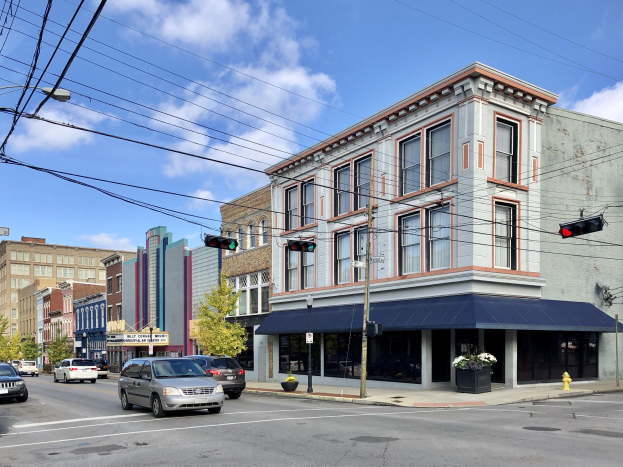 Eine Straße in einer Stadt mit parkenden Autos, Strommästen, Ampeln, Straßenlaternen, Bäumen und Gebäuden unter einem bewölktem Himmel, mit Text, der auf ein kommerzielles Grundstück hinweist, das für $1.250.000 zum Verkauf steht.