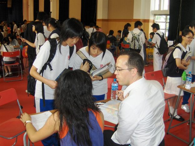 Ein Klassenzimmer mit mehreren Mädchen in weißen T-Shirts, die Bücher halten, vorne, ein Mann und eine Frau, die unten sitzen und diskutieren, und eine gelbe Wand mit einem Glasfenster im Hintergrund.