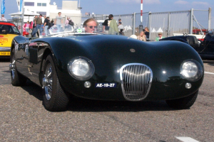 Ein schwarzer Sportwagen auf einem Parkplatz mit einer Person auf dem Fahrersitz, Menschen in der Nähe, einem Zaun, Pfählen, einem Gebäude und einem bewölkten Himmel im Hintergrund.