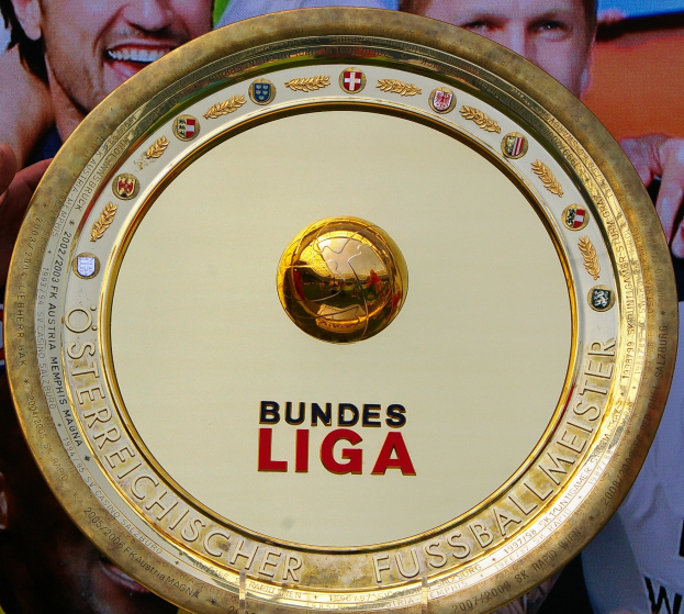Ein Mann hält einen goldenen Schild mit der Aufschrift "Bundesliga" in die Höhe, im Hintergrund ist ein Banner mit drei Personen zu sehen.