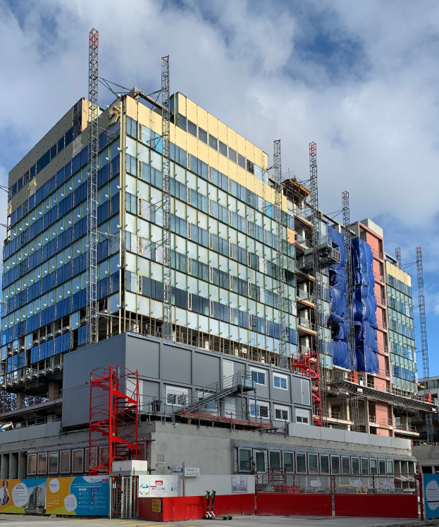 Großes Gebäude im Bau in einer städtischen Umgebung, mit Glasfenstern, Metallrahmen, einer Treppe, einem Zaun, einem Schild, Polen, einem Straßenmast, umgeben von Bäumen und einem bewölkten Himmel.