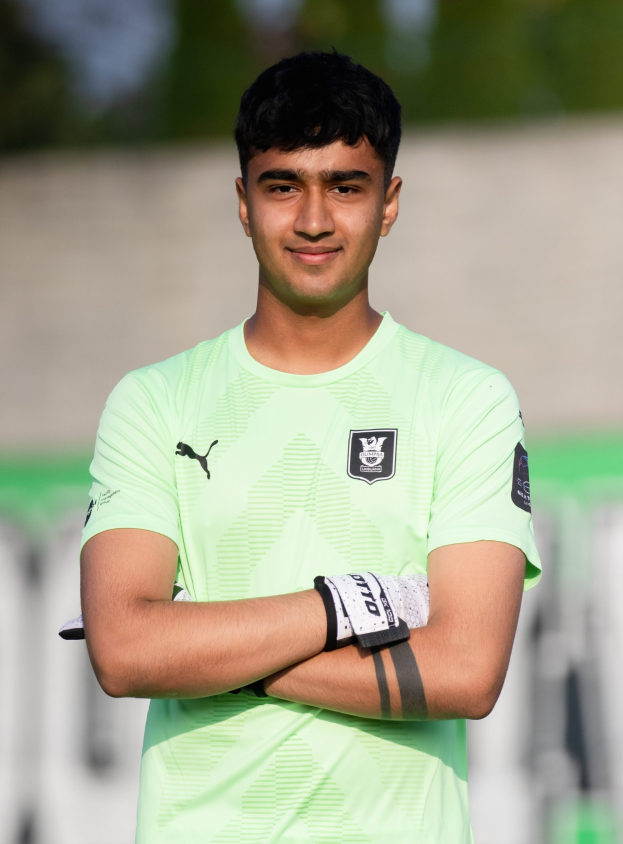 Ein junger Mann in einem grünen T-Shirt steht auf einem Fußballfeld und hat die Arme verschränkt, während er lächelt, mit einem unscharfen Hintergrund.