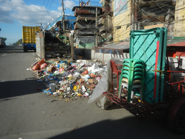 Ein Lastwagen neben einem Haufen Müll auf einer Straße, mit einem Handwagen mit Plastikstühlen rechts daneben und Gebäuden, Strommasten, Bäumen und einem bewölkten Himmel im Hintergrund.