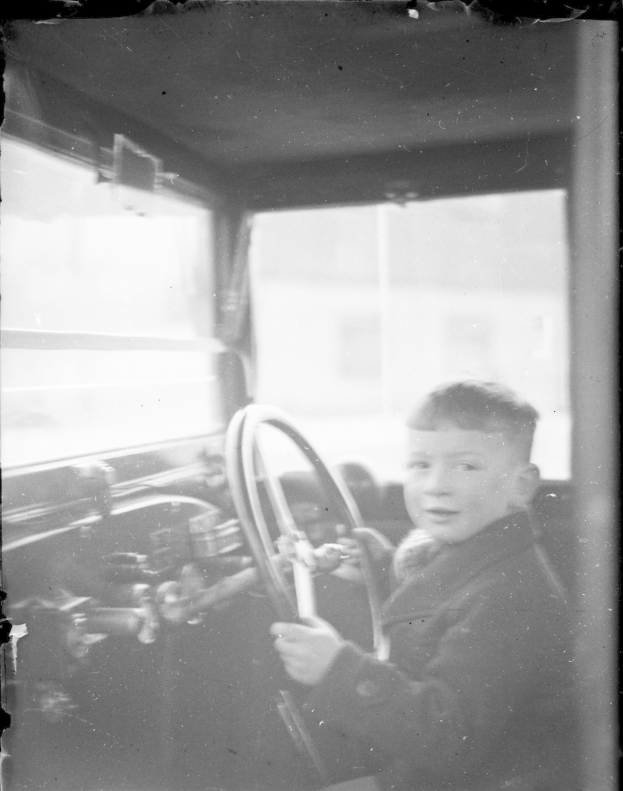 Ein junger Junge in Schwarz und Weiß sitzt im Fahrersitz eines Autos und hält das Lenkrad mit konzentrierter Miene fest.