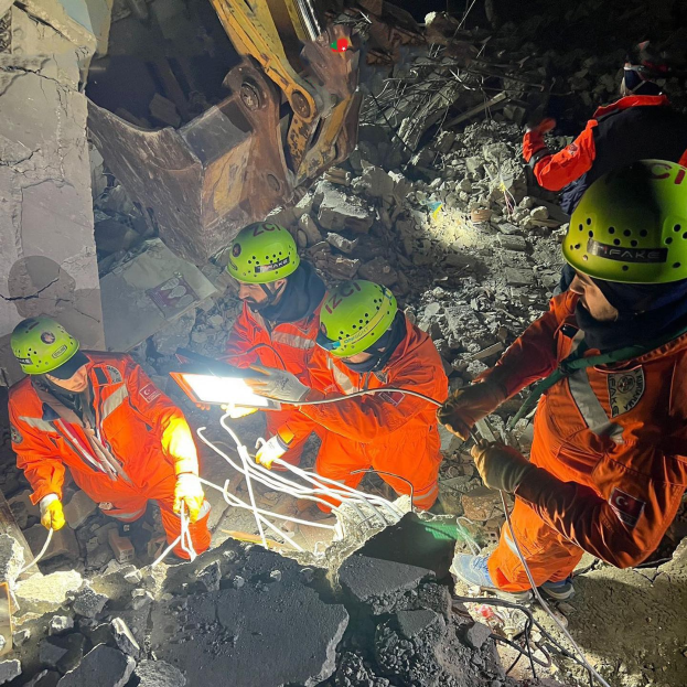 Rettungskräfte in orangefarbener Uniform und grüner Helm entfernen Trömmerschutt von einem eingestürzten Gebäude mit Werkzeugen und Materialien, mit einem Kran im Hintergrund.