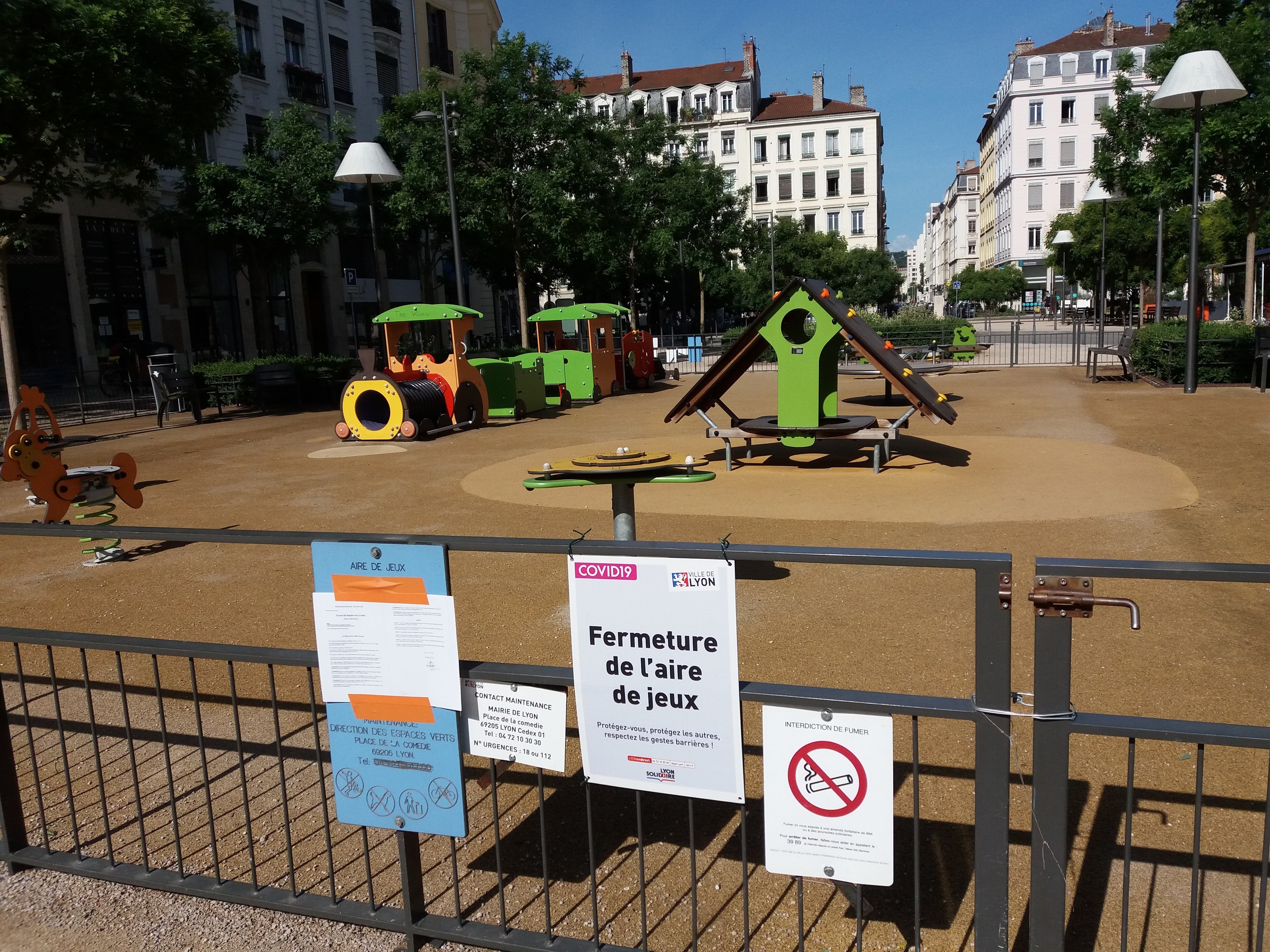 Ein umzäunter Spielplatz in einem städtischen Platz mit Schildern an der Umzäunung, umgeben von Bäumen, Laternenmasten, Gebäuden und einem sichtbaren Himmel.