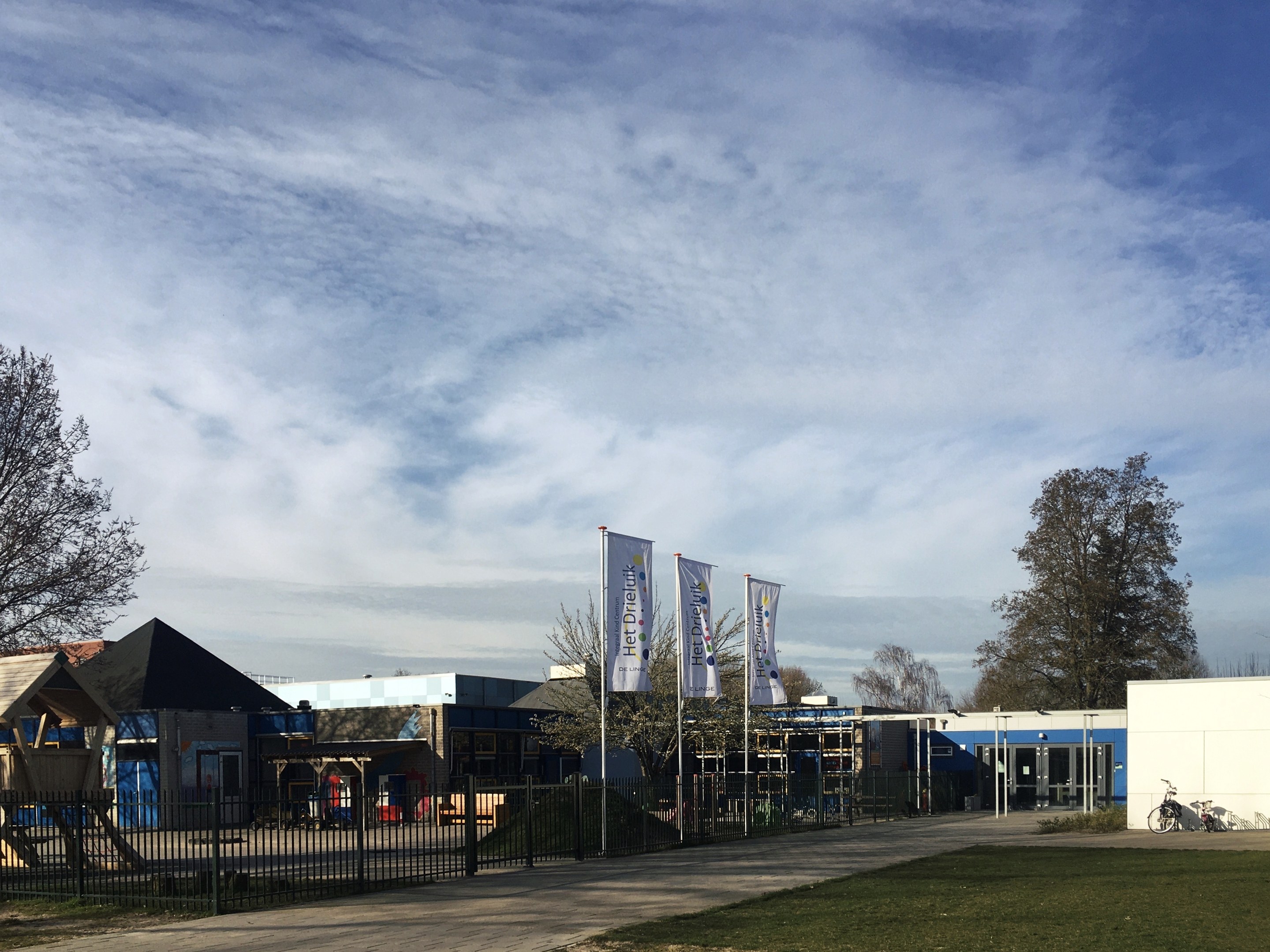 Großes Gebäude mit einem grasigen Bereich davor, umgeben von einem Metallzaun, mit einem Weg, Bäumen, Pfählen, Bannern, einem geparkten Fahrrad, nahen Häusern und einem bewölkten Himmel, als neuer Spielbereich für Kinder an der Schule identifiziert.