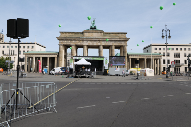 Das Brandenburgertor in Berlin, Deutschland, mit seinen ikonischen Säulen und Statuen, umgeben von einer festlichen Atmosphäre für den Berlin-Marathon, mit Gebäuden, Laternenmasten, Verkehrssignalen, Zelten, Fahrzeugen, Menschen, Fahrrädern und grünen Ballons vor einem bewölkten Himmel.