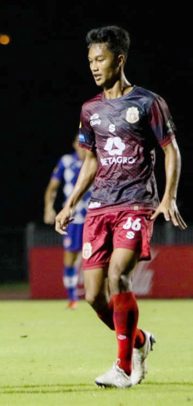 Ein Mann in einem marinefarbenen Hemd und roten Shorts, der Fußball auf einem Feld spielt, mit einer anderen Person im Hintergrund und einer Tafel mit Schrift.