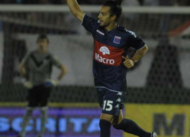 Ein Fußballspieler feiert auf dem Boden nach einem Tor, mit jubelnden Zuschauern und einer Person im Hintergrund sowie einer Texttafel dahinter, alles vor einem leicht unscharfen Hintergrund.