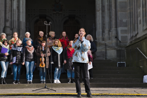 Eine Gruppe von Menschen, einige mit Musikinstrumenten, steht vor einem Gebäude mit Treppen und Geländern, während eine Frau in der Mitte in ein Mikrofon singt, das von Mikrofonstativen umgeben ist.