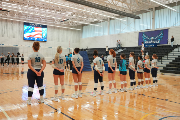 Gruppe von Frauen-Volleyballspielerinnen auf einem Feld mit einem Netz, Treppe rechts, Wand im Hintergrund, Bildschirm oben und Deckenbeleuchtung, bereit für den Beginn eines Spiels.
