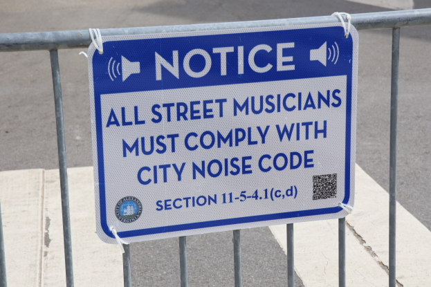 Anschlagtafel an einem Zaun mit einem Schild, das Straßenmusikern die Einhaltung der städtischen Lärmvorschriften auferlegt, mit einer Straße im Hintergrund.