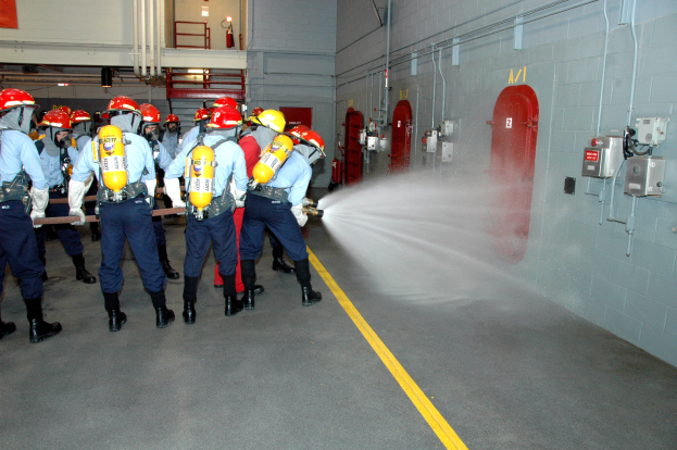 Eine Gruppe von Feuerwehrleuten in Helmen und Zylindern sprüht sich gegenseitig mit Wasser nass, mit Rohren, Feuerlöschern und einer Leiter im Hintergrund.