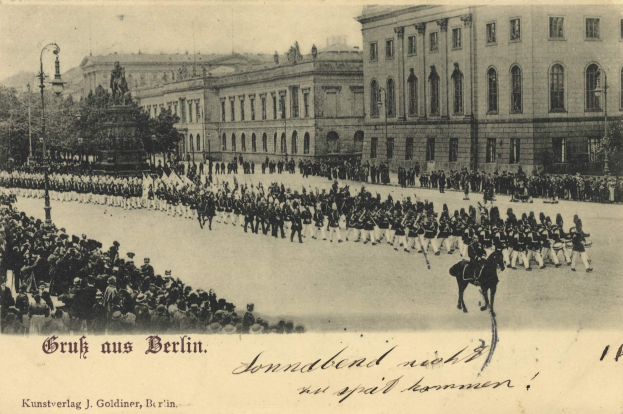Ein altes Schwarz-Weiß-Foto eines Umzugs in Berlin, Deutschland, das Menschen auf dem Boden und auf Pferden, eine Statue, Straßenlaternen, Bäume, Gebäude mit Fenstern und einen bewölkten Himmel zeigt, mit Text am unteren Rand des Bildes.