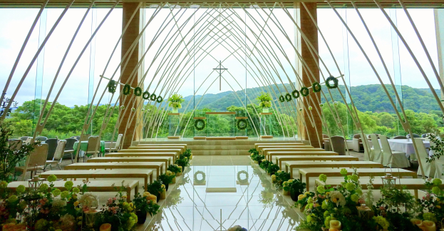 Eine Hochzeitszeremonie in einem großen Saal mit Tischen in einem Halbkreis, dekoriert mit Blumen, unter einem klaren blauen Himmel mit Bäumen und Hügeln im Hintergrund.