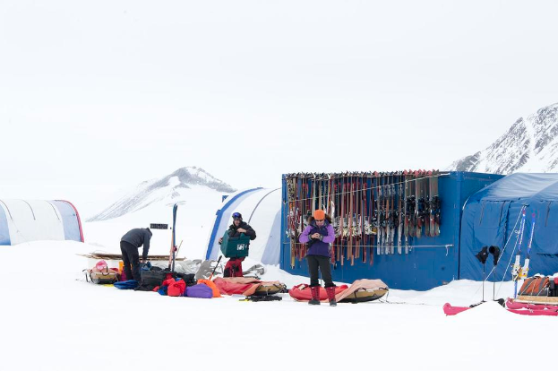 Drei Personen auf einer schneebedeckten Landschaft mit verstreuten Taschen, Zelten mit Skiern im Hintergrund und schneebedeckten Hügeln unter einem klaren Himmel.