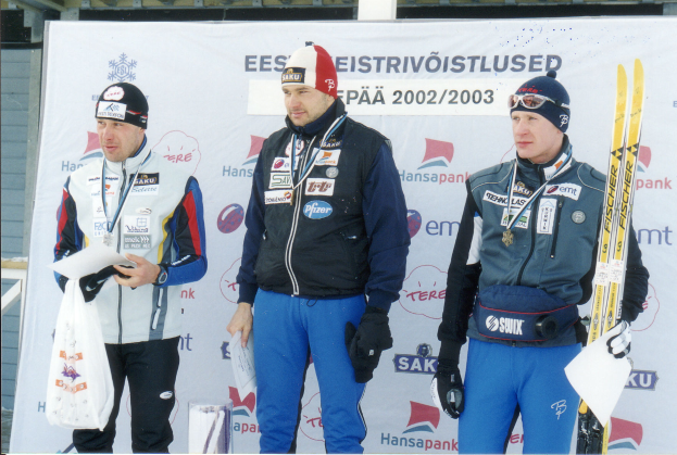 Drei Männer in Ski-Ausrüstung stehen auf einem Podium, jeder hält Ski, Papiere und eine Abdeckung, mit einem Banner im Hintergrund.