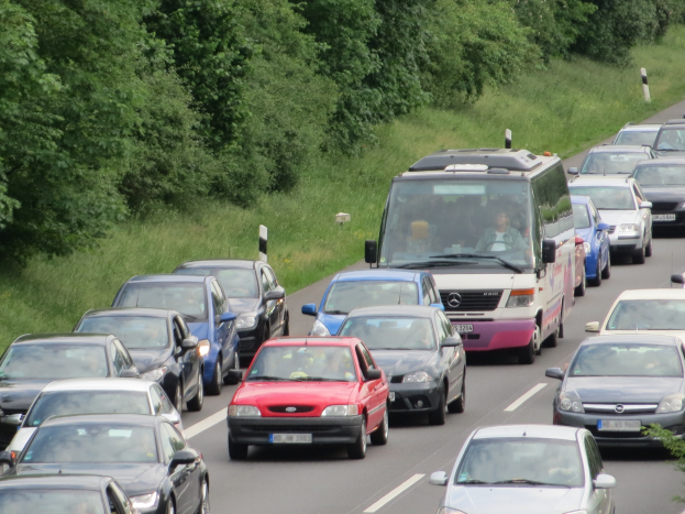 Stau auf einer Autobahn mit vielen Autos, einem Lieferwagen und Menschen in den Fahrzeugen, mit Bäumen und Gras im Hintergrund.