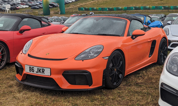 Ein Porsche 718 Boxster GTS beim Goodwood Festival of Speed, umgeben von anderen Autos auf dem Rasen, mit einem Banner, Bäumen und einem klaren blauen Himmel im Hintergrund.