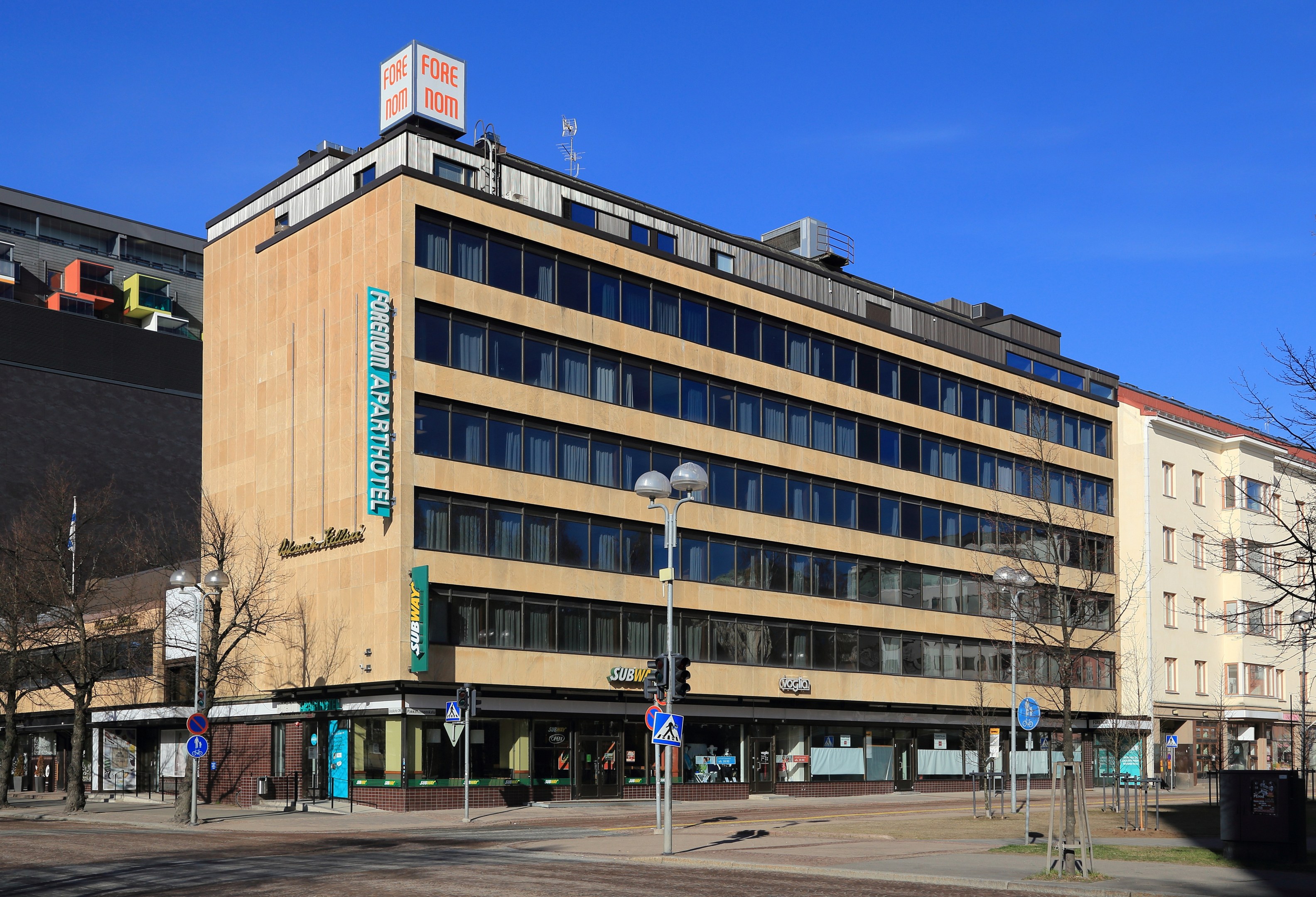 Großes Gebäude mit zahlreichen Fenstern, identifiziert als das Fore Inn Hotel Berlin, umgeben von Straßeninfrastruktur einschließlich Pfählen, Laternen, Signalen, Schildern, Bäumen und einem bewölkten Himmel.