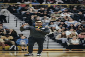 Ein Mann in einer Mütze hält ein Mikrofon und steht auf einem Basketballfeld, hinter ihm sitzen Menschen auf einer Treppe und Taschen liegen auf dem Boden.