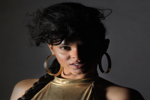 Eine Frau in einem goldenen Kleid und großen Ohrringen steht selbstbewusst vor einer Wand, ihre Haare sind zu einem glatten Bob geschnitten, sie trägt dezentes Make-up mit rosa Wangen und glänzenden Lippen und schaut direkt in die Kamera mit einem entschlossenen Gesichtsausdruck.