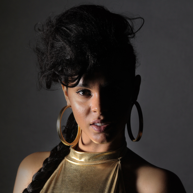 Eine Frau in einem goldenen Kleid und großen Ohrringen steht selbstbewusst vor einer Wand, ihre Haare sind zu einem glatten Bob geschnitten, sie trägt dezentes Make-up mit rosa Wangen und glänzenden Lippen und schaut direkt in die Kamera mit einem entschlossenen Gesichtsausdruck.