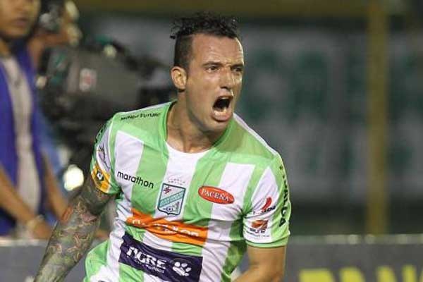 Ein Fussballspieler in grün-weissem Trikot mit Tattoos auf dem Arm feiert nach einem Tor, mit unscharfen Figuren und einer Tafel im Hintergrund.