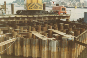 Baustelle mit einem Kran, der Stahlstangen von einem großen Haufen hebt, umgeben von Wasser, Gebäuden, Bäumen und einem klaren blauen Himmel.