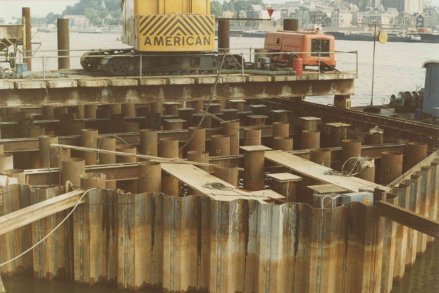 Baustelle mit einem Kran, der Stahlstangen von einem großen Haufen hebt, umgeben von Wasser, Gebäuden, Bäumen und einem klaren blauen Himmel.