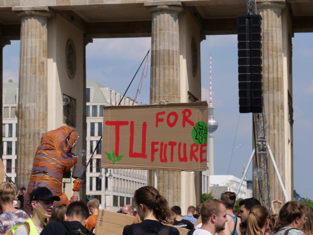 Gruppe von Menschen vor dem Brandenburger Tor in Berlin, Deutschland, mit einigen Taschen tragend, einer Person in Kostüm mit einer Tafel und Gebäuden, Bäumen und einem Turm im Hintergrund unter einem bewölkten Himmel.