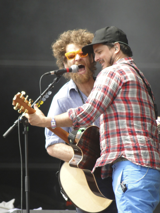 Zwei Männer mit Gitarren und Mikrofonen auf einer Bühne, einer trägt ein rot-blau kariertes Hemd mit blauen Hosen, der andere eine schwarze Mütze und eine Schutzbrille.