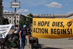 Eine Gruppe von Menschen steht vor einem Gebäude und hält ein gelbes Banner hoch, auf dem "Hope Dies, Action Begins" steht. In der Nähe stehen Fahrräder und ein Mast mit einer Tafel. Im Hintergrund sind Bäume und ein klarer blauer Himmel zu sehen.