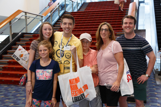 Eine Gruppe von Menschen steht zusammen vor einer Rolltreppe, lächelt und hält Taschen, bei der Nationalen Buchmesse 2019, mit Treppen, Geländern und einer Wand im Hintergrund.