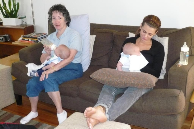 Zwei Frauen auf einem Sofa sitzend, die Babys halten, mit Kissen auf dem Sofa, einer weißen Wand im Hintergrund und einem Schreibtisch mit Büchern und einer Zimmerpflanze links.