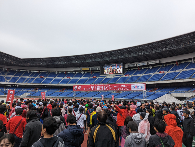 Eine große Menschenmenge steht vor einem Stadion während der Eröffnungszeremonie der Olympischen Spiele Tokio 2020, mit Bannern, Stühlen und einem Bildschirm im Hintergrund, unter einem sichtbaren Himmel und einem Wasserzeichen in der rechten unteren Ecke.