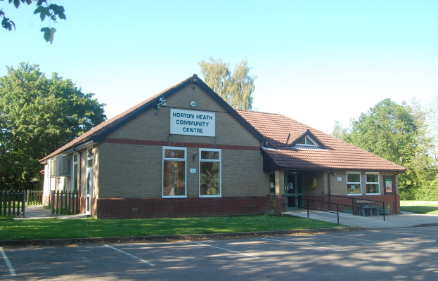 Ein Haus mit einem Dach, Fenstern, einem Schild mit der Aufschrift "Horton Heath Community Centre", einem Metallzaun, einer Bank, Pflanzen, Gras, einem Weg, einer Gruppe von Bäumen und einem bewölkten Himmel.