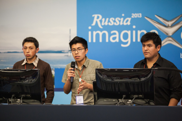 Drei Jungen stehen neben einem Schreibtisch mit zwei Computersystemen, einer spricht in ein Mikrofon und trägt eine ID-Karte mit rotem Tag, vor einer Wand mit einer Werbetafel, auf der "Russia Imagine 2013" steht.