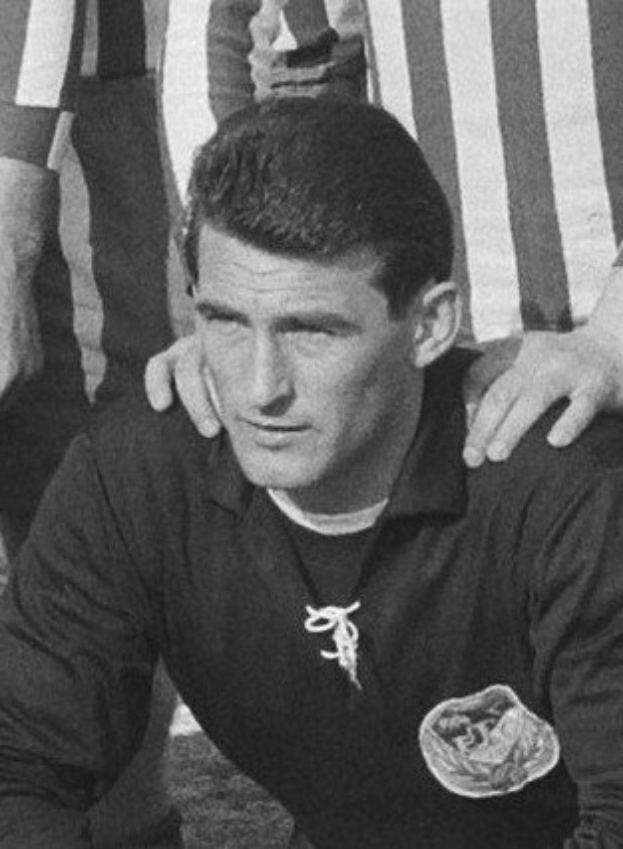 Schwarzes und weißes Foto eines Mannes, der auf einem grünen Fußballfeld sitzt und ein T-Shirt und eine Kette trägt, mit zwei stehenden Figuren hinter ihm.