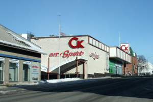 Ein Sportgeschäft in einer Stadt an einer Straßenecke mit umliegenden Gebäuden, Straßenmöbeln, Bäumen und Schnee unter einem sichtbaren Himmel.