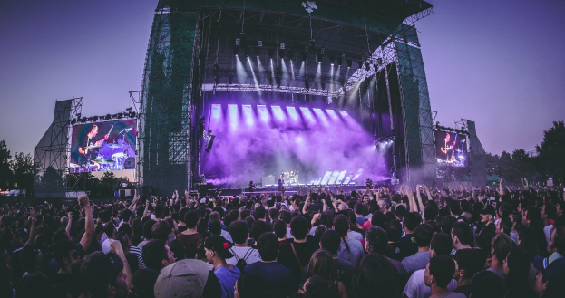 Eine gro├če Menge bei einem Musikfestival schaut auf einer hell erleuchteten Bühne mit Bildschirmen, die vor einem Geb├Ąude, B├Ąumen und einem klaren blauen Himmel steht.