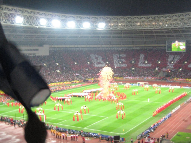 Ein Stadion mit Flutlicht, Eisenstäbe, Zuschauern, stehende Personen, Werbetafeln, Anzeigetafeln, Überwachungskameras und dekorative Fahnen.
