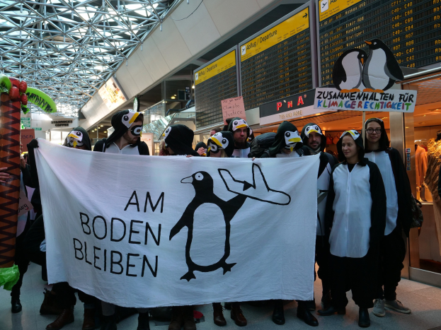 Gruppe von Menschen in Pinguinkostümen mit einem Banner vor einer Wand mit Pinguin-Displays, Schaufensterpuppen in Kleidern und Deckenbeleuchtung.