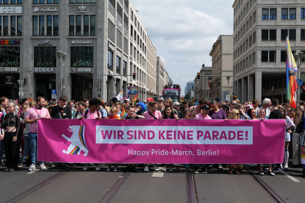 Eine Gruppe von Menschen marschiert auf einer Straße in Berlin, Deutschland, mit einer pinken "Happy Pride March"-Fahne, während Gebäude, Laternenpfähle und Verkehrszeichen die Straße säumen und der Himmel bewölkt ist.