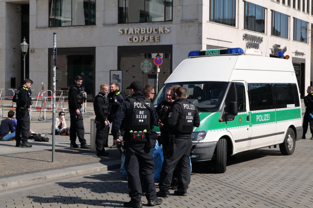Eine Gruppe von Polizisten steht vor einem Starbucks-Café mit einem Van auf der rechten Seite und einigen Menschen auf der linken Seite, während im Hintergrund ein Gebäude mit Fenstern, einem Schild, einem Laternenmast und einem Zaun zu sehen ist.